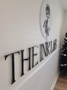Photograph of Custom Acrylic signage - 1/4" with hidden standoffs for mounting. Reads "The Ink Lab" with their rose logo