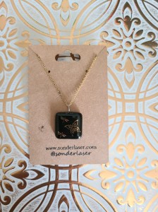 Photo of necklace attached to a cardstock backer for hanging on display. This cardstock backer is stamped with the website and social media handle of the small business. Background is white with gold filigree design.