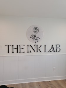 Photograph of Custom Acrylic signage - 1/4" with hidden standoffs for mounting. Reads "The Ink Lab" with their rose logo