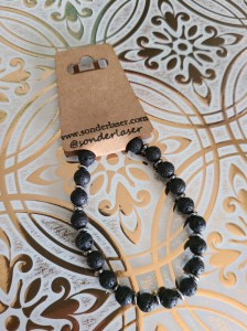 Photo of a bracelet attached to a cardstock backer for hanging on display. This cardstock backer is stamped with the website and social media handle of the small business. Background is white with gold filigree design.