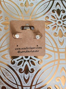 Photo of diamond earrings attached to a cardstock backer for hanging on display. This cardstock backer is stamped with the website and social media handle of the small business. Background is white with gold filigree design.