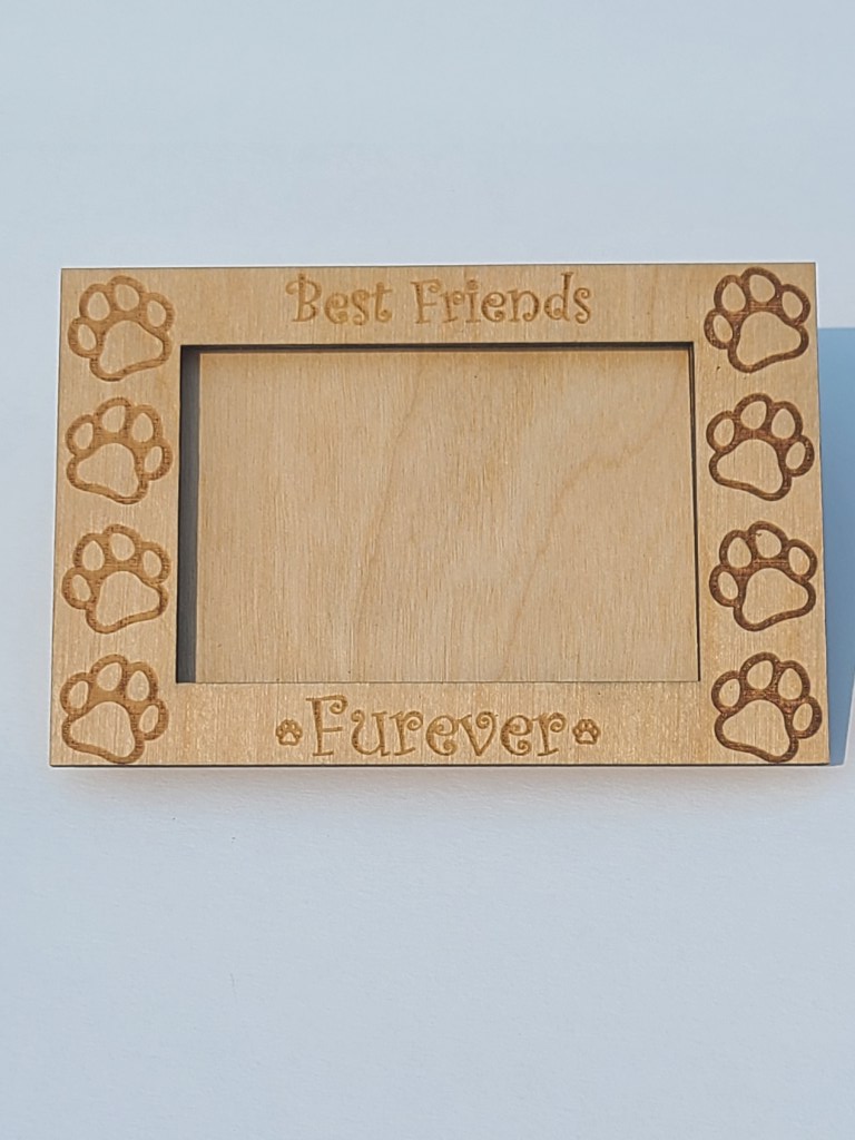 Photograph with a plain white background showing a wooden magnetic picture frame that says "Best Friends Furever" with paw prints engraved around the edges.