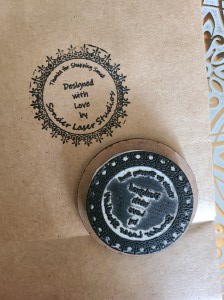 Photograph of stamp filled with ink laying on brown paper bag that has just been stamped. Custom stamp reads, "Thanks for shopping small. Designed with love by Sonder Laser Studios."