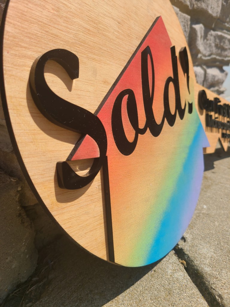 Photograph of wooden key sign showing a house painted in rainbow and the word "Sold" across it in black acrylic.