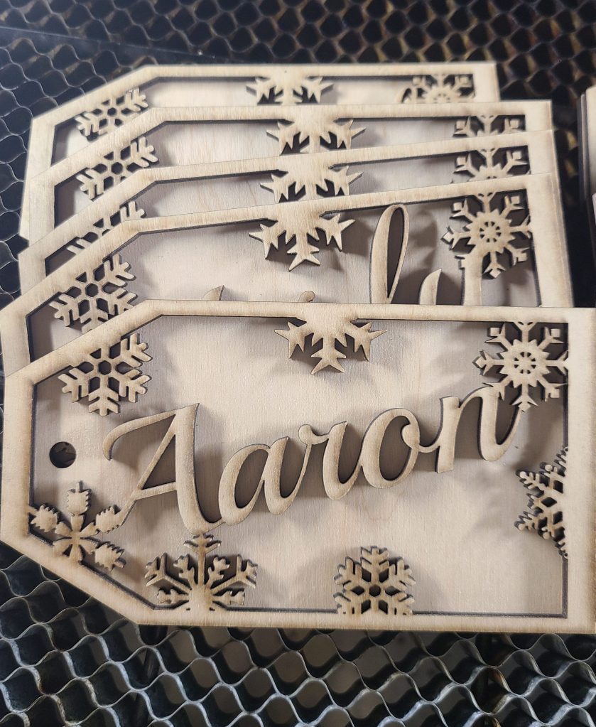 Photograph of two-layer wooden gift tags. The front layer displays a name and snowflakes, back layer is solid. 