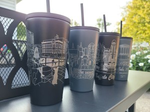 Photograph of City of Good Neighbors: The Collection - stainless steel tumblers, set of four. Pictured outside with trees and flowers in the background