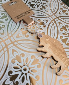 Photo of Buffalo shaped wooden keychain with beach scene engraved inside of it. Keychain is attached to a cardstock backer for hanging on display. This is stamped with the website and social media handle of the small business. Background is white with gold filigree design.