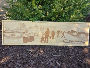 Photograph of a wooden sign displaying a vibrant scene captures the lively atmosphere of Canalside, where people glide across the ice in the foreground. Featuring Shark Girl, the NFTA Metro train car, and visitors enjoying the area, the scene also includes the Explore ‘n More Children’s Museum, complete with its giant tricycle sculpture on the roof. In the distance, the USS The Sullivan, a historic naval ship, proudly rests at the Buffalo and Erie County Naval & Military Park after being successfully righted following a hull incident. The iconic Skyway and nearby hotel provide a stunning backdrop, embodying the essence of Buffalo’s waterfront experience.