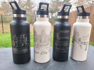Photograph of four stainless steel tumblers in an outdoor setting. Photo features engraved tumblers with four different scenes from Buffalo, NY. Left to Right: Theatre District, Buffalo City Hall, Art Gallery, and Canalside.