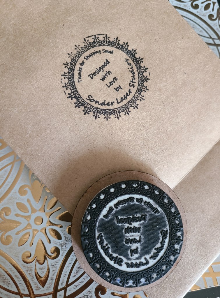 Photograph of stamp filled with ink laying on brown paper bag that has just been stamped. Custom stamp reads, "Thanks for shopping small. Designed with love by Sonder Laser Studios."