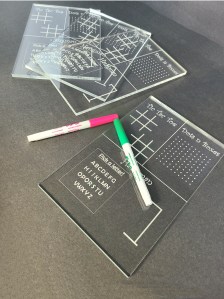 Photograph of Clear acrylic dry-erase game boards featuring three classic games - tic tac toe, dots n boxes, and hangman. Photograph also shows two dry erase markers.