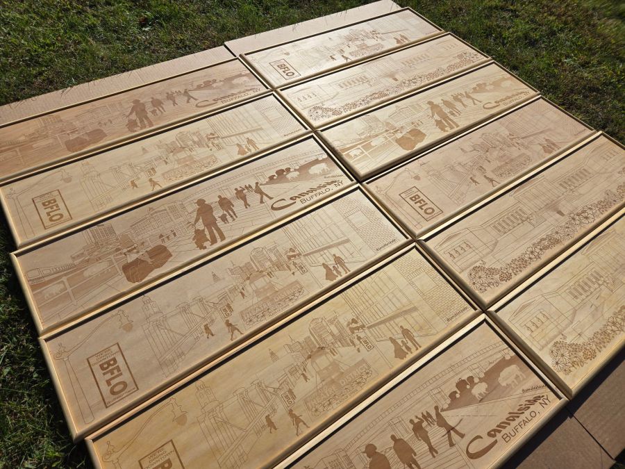 An array of custom wooden signs showcasing intricate laser-engraved scenes of Buffalo, NY, lying in the sun on lush green grass. The signs display vibrant designs representing iconic landmarks, with each piece framed in polished wood, reflecting the craftsmanship and artistry behind their creation.